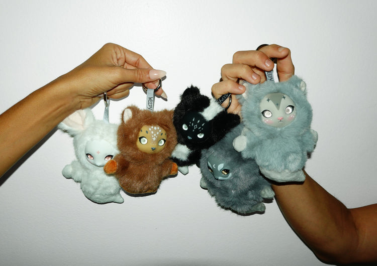 Two hands holding all five Forest Spirits plush charms on keyrings against a white wall