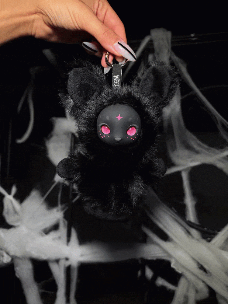 A hand holding Luma, the black bunny Nightfall plush charm with pink eyes, against white cobwebs