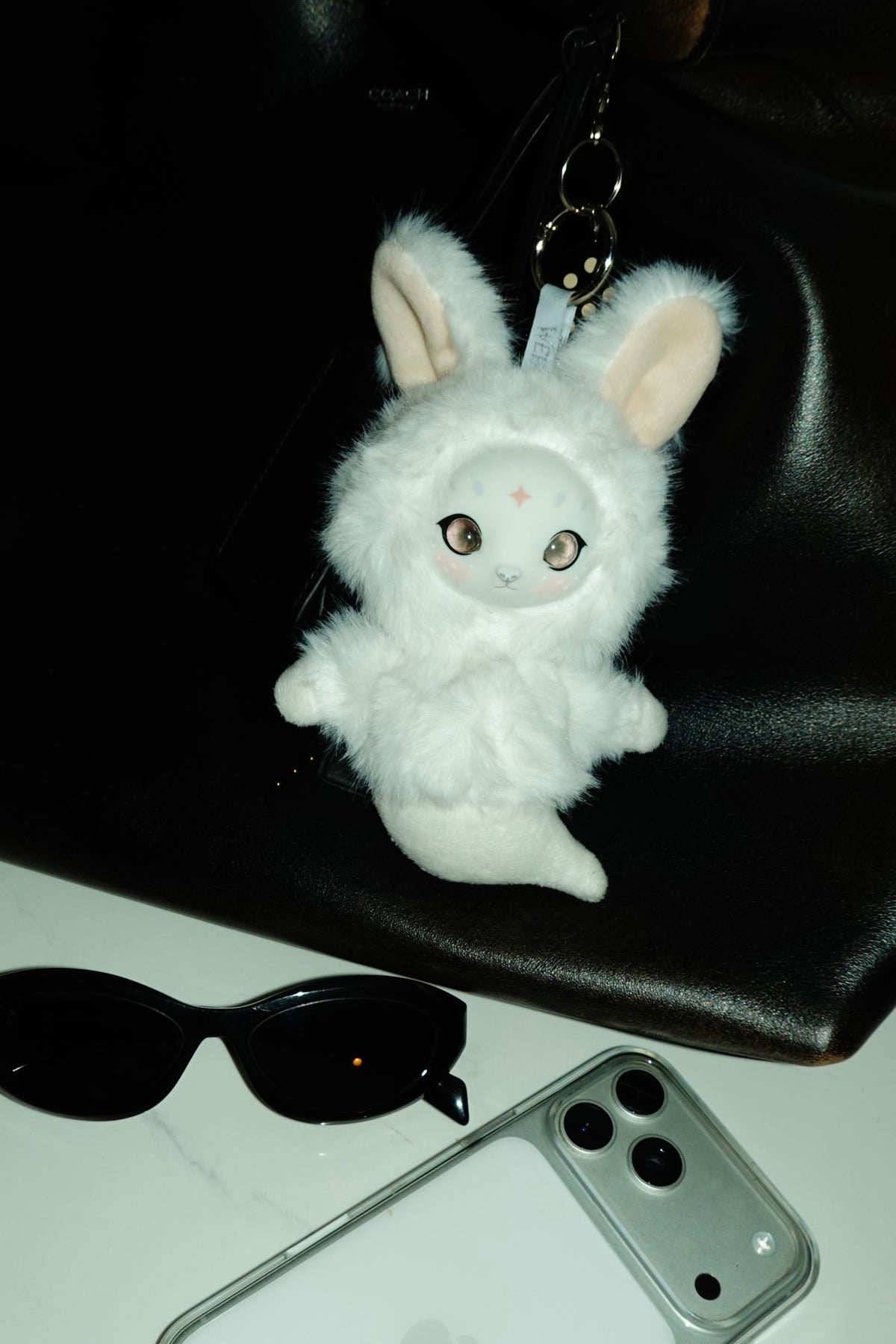 White fluffy rabbit keychain with large ears on a black back, next to sunglasses and a phone.