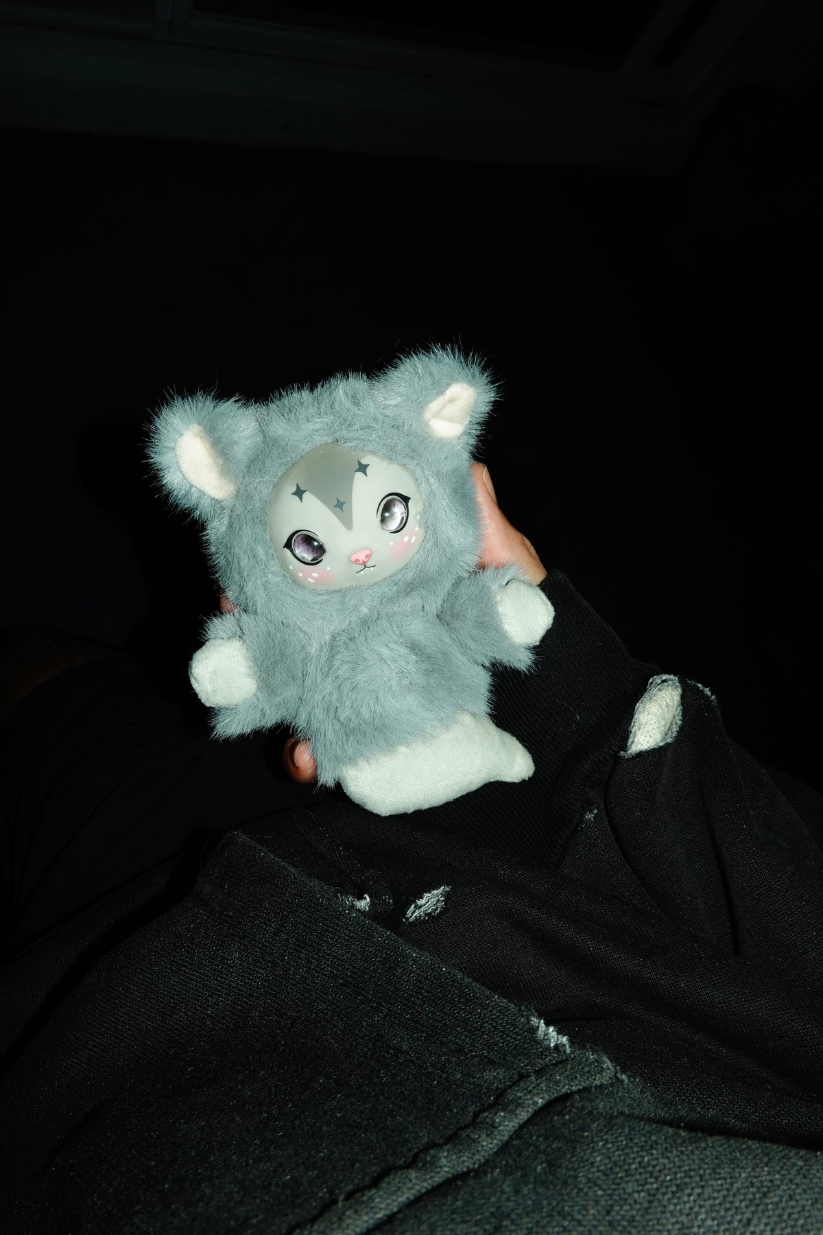 Gray opossum plush charm with white accents held by a person against a dark background