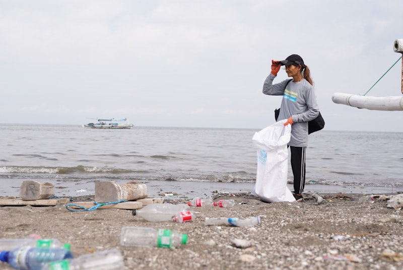 Person cleaning up trash on a beach with bottles and bags scattered around.
