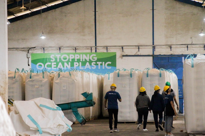People walking through a recycling warehouse with 'Stopping Ocean Plastic' sign and large bags.